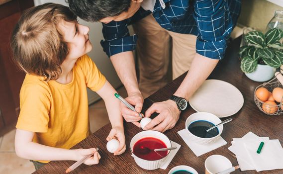 Easter day. Father and son painting eggs on wooden background. Family sitting in a kitchen. Preparing for Easter, creative homemade decoration.