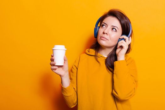 Happy person with cup of coffee listening to music on headset. Caucasian woman feeling relaxed and enjoying song on headphones while holding hot beverage to drink. Young adult relaxing