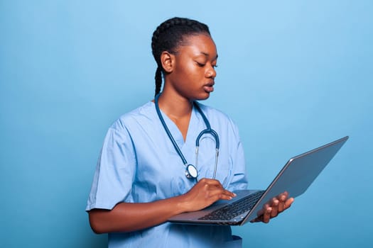 African american specialist assistant holding laptop computer typing disease expertise working at medical treatment in studio with blue background. Physician woman in uniform. Health care service