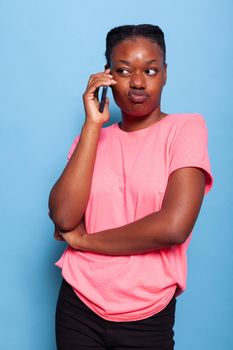 Portrait of dissatisfied african american young woman talking at phone with friend sharing rumors while standing in studio with blue background. Communication concept. Remote connection
