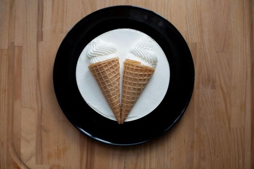 round white dessert sweet tasty ice cream cake with decoration two ice cream cones in a waffle cup on a black plate on a wooden board background. top view, close-up