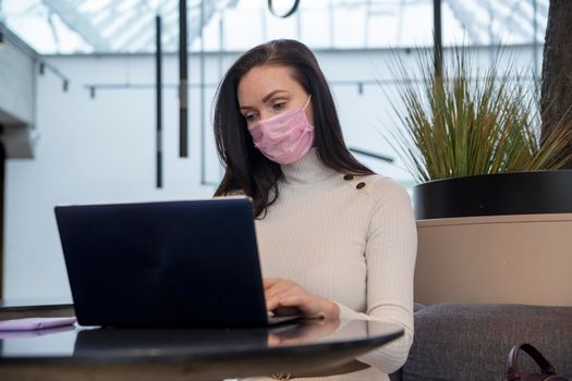 young woman with protective face mask works on laptop in cafe.