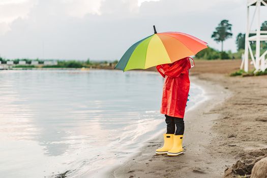 Boy in a red raincoat and yellow rubber boots stands at river bank and holding rainbow umbrella. School kid standing still near autumn lake. Child wearing waterproof clothes at shoreside.