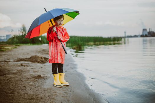 Boy in a red raincoat and yellow rubber boots stands at river bank and holding rainbow umbrella. School kid standing still near autumn lake. Child wearing waterproof clothes at shoreside.