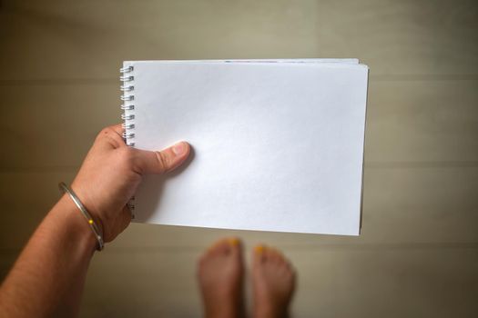 top view of a hand with silver bracelet holds a white empty paper notepad with copyspace,creative concept, close up, in the background is a wooden floor and feet in a blur.