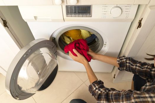 A girl folds bright clothes into a new washing machine in the bathroom at home, a woman washes things and spin them out with detergent in the laundry close-up, top view.