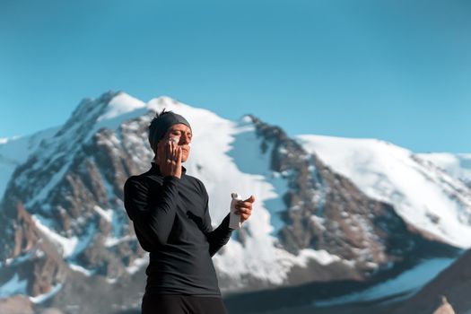 A young man applies sunscreen to his face in the highlands, in the background the peaks of the mountains are covered with snow. The traveler takes care of his skin while climbing.