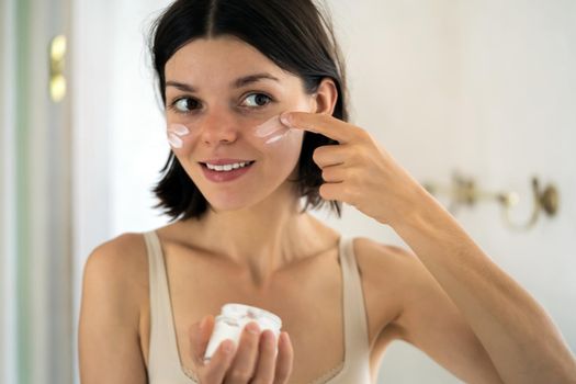 A young girl applies a moisturizing cream mask near the mirror in the bathroom and holds a jar of balm in her hand. A woman takes care of the health of her skin, uses cosmetic masks.