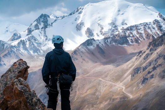 A young man traveler is engaged in mountaineering. Hiker in a helmet, with a rope climbs to the top, against the backdrop of a stunning view with snow-capped mountains.