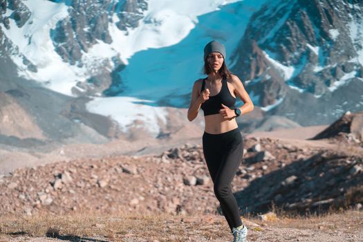 A young athletic girl jogging in a scenic area among the snow-capped mountains at sunset. A runner in leggings and a top is warming up, exercising, and involved in trail running.