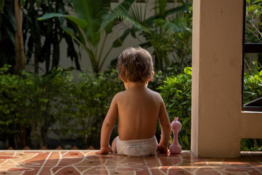 a charming toddler sits with a toy on the steps of a house in the sunlight. back view.