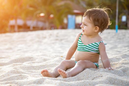 happy toddler in a swimsuit sits on a sandy beach in the sunshine