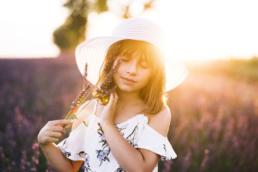 Close up portrait of young happy little girl with a hat on her head in lavender fields on the background. Kid girl dressed in a hat in spring time.