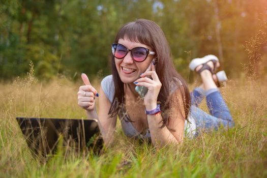 young woman lies on the grass with a laptop and speaks emotionally on the phone