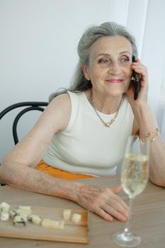 Beautiful old grandmother with grey hair and face with wrinkles is using smartphone, talking with someone and sitting at the table at home on window background, mother's day, happy retirement.