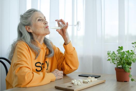 Beautiful old grandmother with grey hair and face with wrinkles sitting at the table at home on window background with glass of champagne, mother's day, happy retirement.