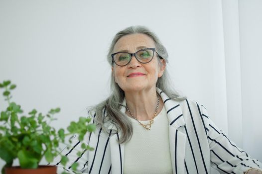 Senior grey haired businesswoman in striped jacket with eyeglasses is working in her office sitting at the desk. Happy retirement, employment and labor.