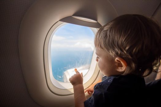 Toddler looks at the ground through the porthole of a flying plane. travel with children