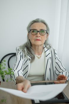 Senior grey haired businesswoman in striped jacket with eyeglasses is working with documents in here office sitting at the desk. Happy retirement, employment and labor.