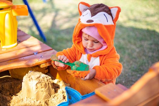cute toddler in orange jumpsuit plays in the sand in the sandbox outdoors on a sunny day