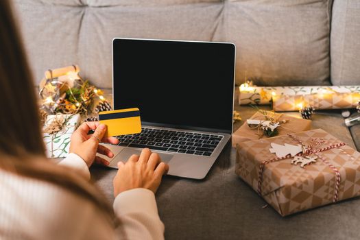 Close up of woman hands with credit card, gifts, and mock up screen laptop. Online shopping at Christmas holidays. Cropped female sit on couch with natural eco presents and decor. Merry Christmas.