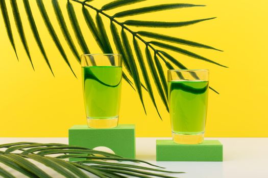 Creative composition with sambuca, liqueur or green alcohol drink in shot glasses on geometric shapes against yellow background with palm leaf. Concept of summer parties and alcohol cocktails