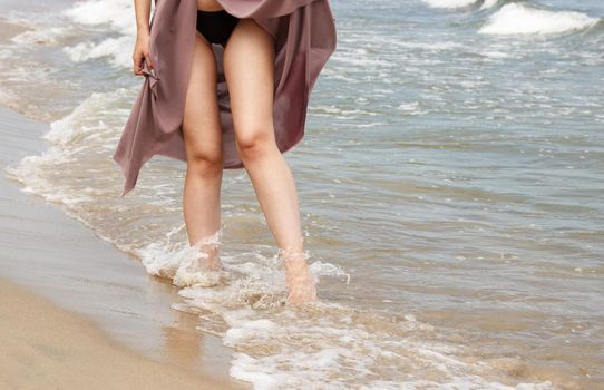 legs of a young woman in black bikini and beige dress standing on sand by sea on sunny summer day