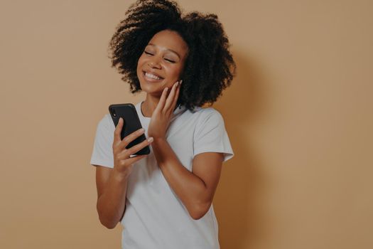 Lovely young african american woman with modern smartphone in hands feeling happy to get message from her boyfriend, looking at device screen and smiling while standing over beige studio wall