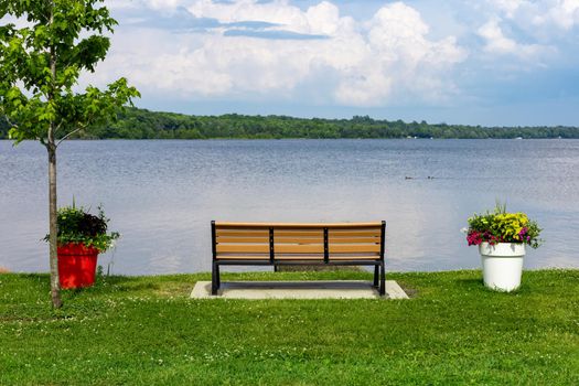 A park bench and large flowerpots are on the shore of the Ottawa River in a public park. The bench faces the water, where ducks swim in the distance on a cloudy day.