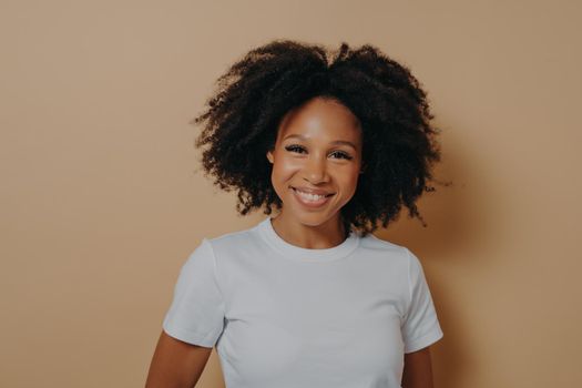 Beautiful cheerful dark skinned woman feeling happy, smiling at camera, carafree young african female in white tshirt expressing positivity while posing against beige background. Happiness concept