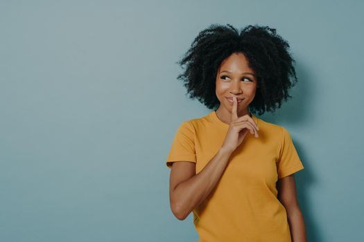 Keep silence gesture. Cheerful afro girl showing shhh sign with finger near lips, standing over pastel blue background with copy space and mysteriously looking away. Positive women emotions concept