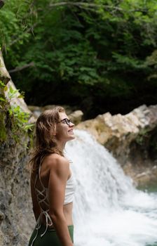 Summer travel. Young happy woman enjoing the waterfall. Woman standing in front of waterfall and smiling