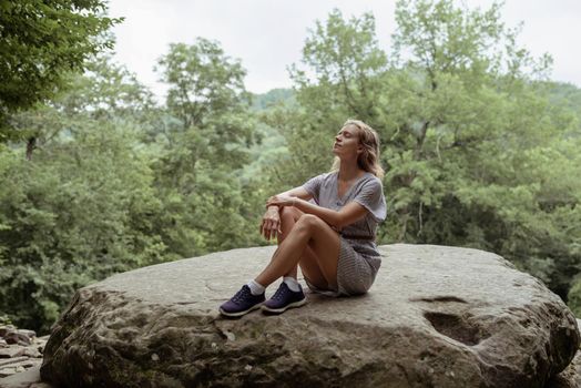 Travel concept. Young woman in summer dress sitting on a big rock in the forest, having rest or meditating