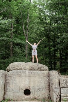 Travel and freedom concept. Back view of a young woman in summer dress standing on a big rock in the forest looking away