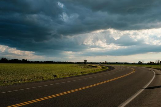 A two-lane road curves into the distance as it passes by farm fields under dramatic clouds of a coming summer storm.