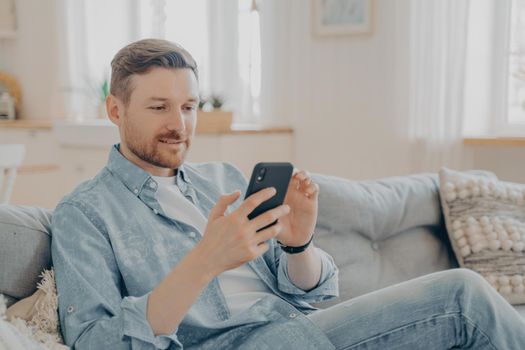 Young man in casual clothes surfing web on his phone and relaxing, checking messages and notifications while sitting on comfortable couch with legs crossed, blurred background