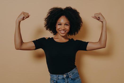 Young confident african lady in casual clothes showing strength of her arms by lifting them up, isolated over dark beige studio wall with copy space. Positive women emotions and body language concept
