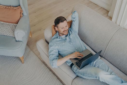 Happy young business company worker in casual clothes relaxing and smiling with confidence while lying on couch with hand behind head after successful online presentation