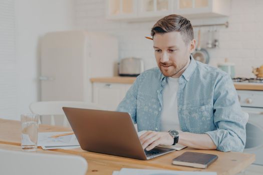 Focused young man freelancer using laptop while working from home at his workplace, millennial guy typing on pc notebook and surfing in internet, looking at screen while sitting at kitchen table