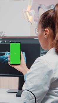 Woman looking at green screen on mobile phone for stomatological care at dentistry office. Orthodontist holding smartphone with chroma key and mockup template on display for teethcare