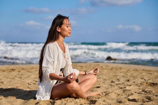Young beautiful woman in lotus position meditating on the beach. Relaxing resting Asian female with closed eyes sitting on sand, scenic seascape background. Lifestyle, health, beauty, nature, people