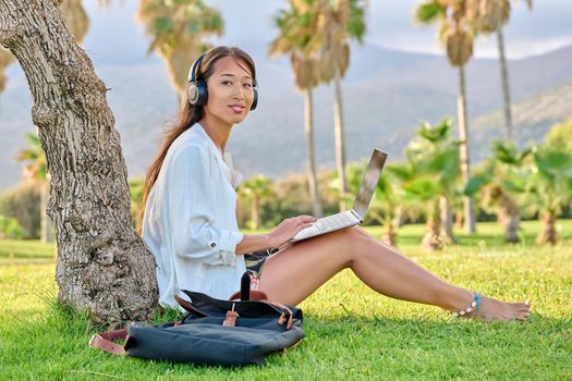 Young woman in headphones with laptop sitting on grass in park. Female student, creative freelancer, businesswoman, blogger working, studying remotely. Education, online business blogging technologies