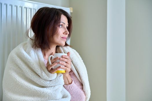 Cold winter autumn season at home, frozen woman basking herself with a warm blanket and a mug of tea near the central heating radiator