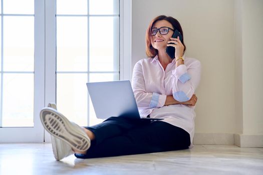 Business woman sitting on floor with laptop talking on smartphone, home office. Female psychologist counselor teacher mentor therapist analyst working online. Technology in business education medicine