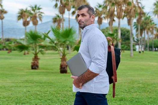 Portrait of serious confident mature male with laptop outdoors. Businessman with backpack, laptop in hands, tropical park nature. Business, technology, freelance, lifestyle, middle age, business trip