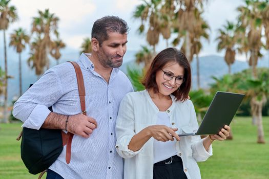 Middle-aged man and woman looking in laptop screen outdoors. Couple of business colleagues talking discussing smiling, tropical park nature background. Business, people, technology, mature age concept