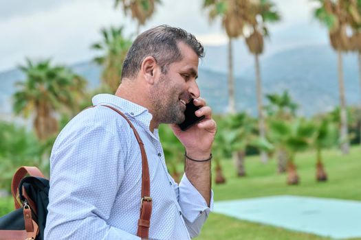 Mature business man talking on the phone outdoors. Serious confident male with backpack, in tropical park. Business trip, freelance, communication, technology, middle aged people concept, copy space