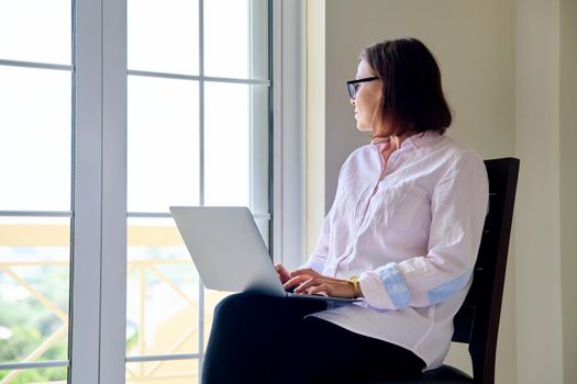 Mature businesswoman in glasses with laptop in her hands near window. Remote work, education, business, freelance, technology concept