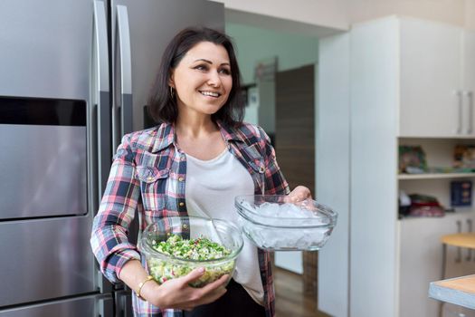 Middle aged woman in the kitchen with salad and a bowl of ice in the kitchen near the refrigerator. Homemade food, lifestyle, home, people concept