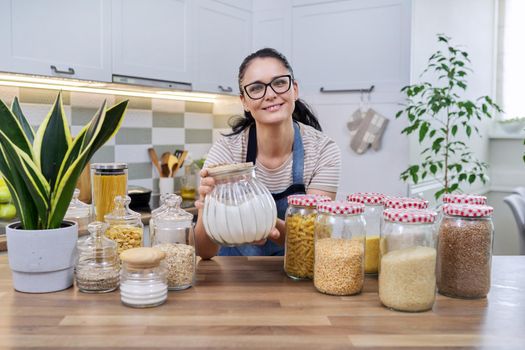 Food, grocery storage, smiling woman looking at the camera in the kitchen, with a can of flour in hand. Organization of storage of bulk products in glass jars and containers, pantry